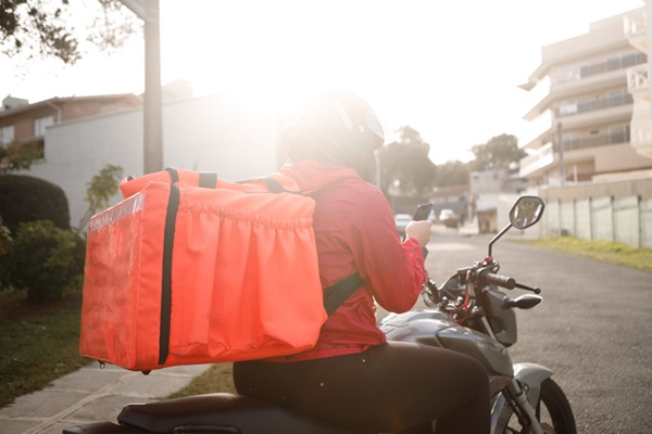 Fotografia de um moto entregador parado com um celular na mão. Destaque na imagem para a caixa de entregas na cor laraja nas costas. O moto entregador está em uma rua com casas ao fundo e o sol se pondo, em contra luz.