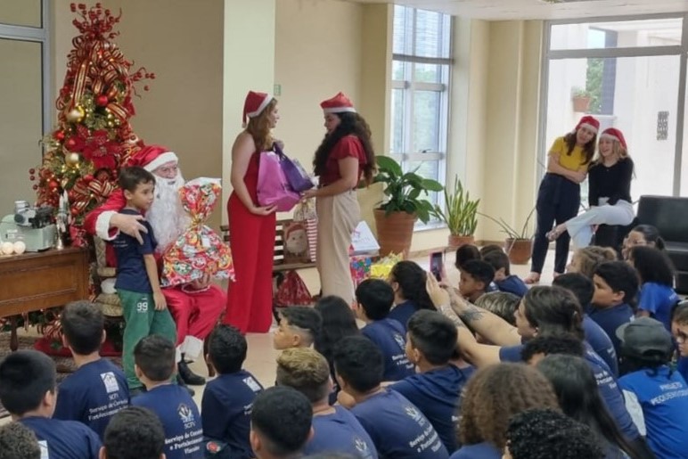 Sala decorada com árvore de natal, Papai Noel sentado, crianças e adultos interagindo, trocando presentes, enquanto servidoras observam o evento animadamente.