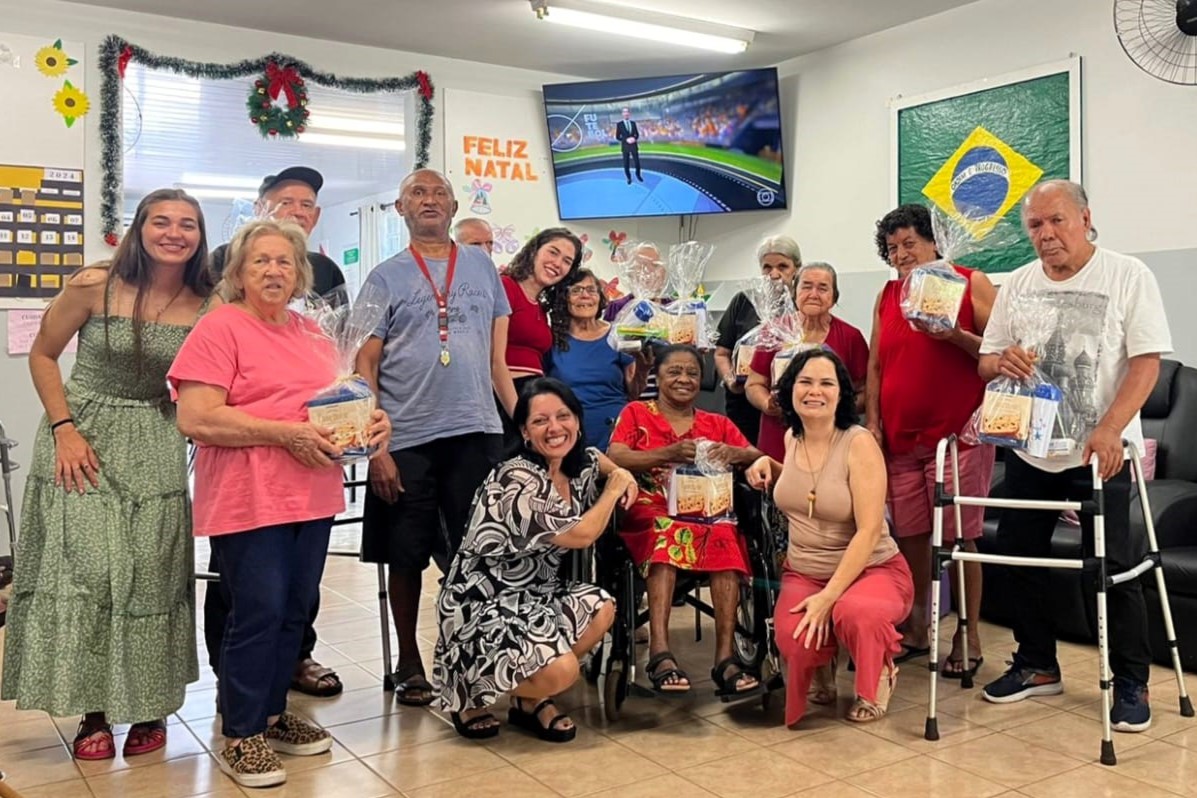 Grupo de pessoas em um ambiente decorado com tema natalino, segurando cestas de presente e sorrindo para a foto, incluindo idosos, cuidadores e uma bandeira do Brasil ao fundo.