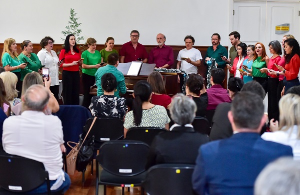 Fotografia do Coral do TRT, composto por 17 pessoas organizadas em formato de U. no centro, um piano sendo tocado pelo maestro. Em primeiro plano, pessoas de costas sentadas em cadeiras assistem a apresentação.