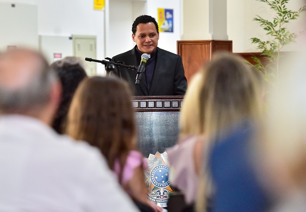 O padre Salles Nogueira, homem de cabelos encaracolados, veste roupa preta e fala em frente a um microfone e uma tribuna. Em primeiro plano, pessoas de costas sentadas em cadeiras o ouvem.