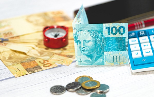 Notas e moedas de reais sobre uma mesa de madeira. Uma calculadora e um relógio na composição