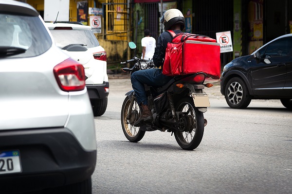 Um entregador de comida de aplicativo é visto andando de moto em meio a carros em avenida de uma cidade brasileira. Ele carrega uma mochila de costas grande e vermelha, típica de entregadores de comida.