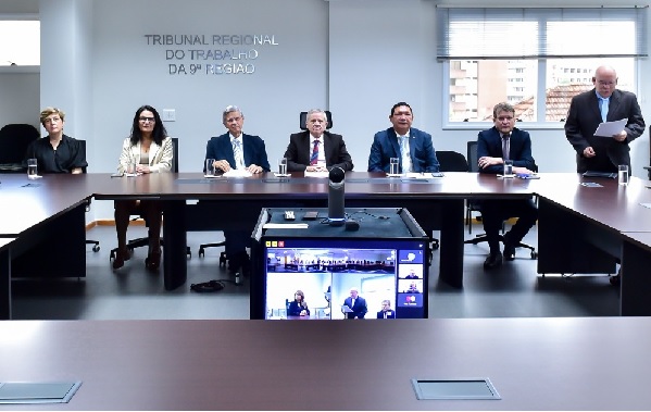 Fotografia da mesa de abertura do Ano Correcional mostra seis pessoas sentadas em frente a uma mesa e uma pessoa em pé. Na frente, uma bancada com uma câmera de vídeo e monitores que mostram os participantes de uma teleconferência.