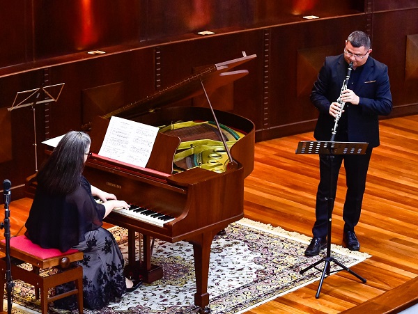 Fotografia a partir do alto mostra uma mulher de costas tocando um piano de cauda e ao lado do instrumento um homem toca clarinete.