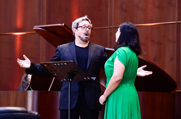 Homem e uma mulher cantam frente a frente um com o outro com um piano de cauda atrás deles. Ela está parcialmente de costas e veste um vestido verde. Ele é um homem branco de cabelos descoloridos e bigode, usa terno escuro com camiseta.