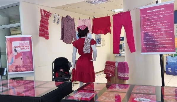 Uma exposição com roupas penduradas em um varal, incluindo um vestido vermelho em um manequim. À direita, um cartaz com a frase 