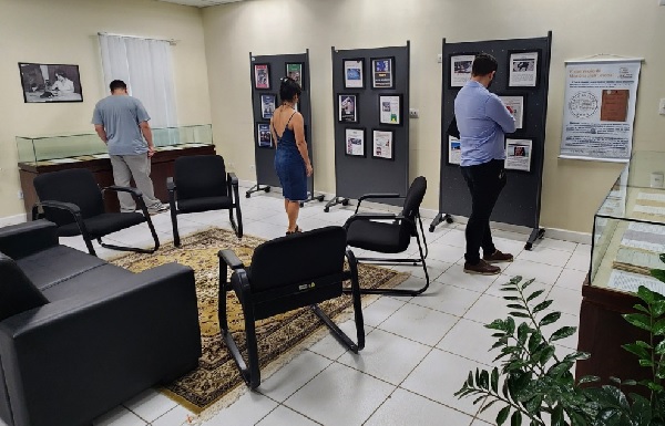 Fotografia de uma sala com sofás e cadeiras e, próximo das paredes, mesas com vidros e paineis com fotografias. Três pessoas, de costas, olham para a exposição, dois homens e uma mulher.