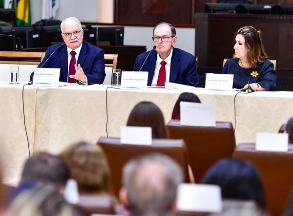 Ministro Edson Fachin fala sentado em frente a uma mesa com toalha creme. Ao lado, o presidente do TRT-PR, desembargador Célio Waldraff, e a diretora da Escola Judicial, desembargadora Ana Carolina Zaina. Em frente deles, uma plateia acompanha o discurso.