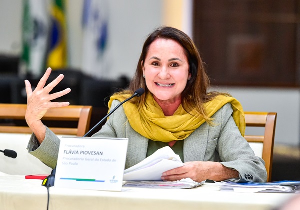 A advogada Flávia Piovesan fala em frente a um microfone sentada em frente a uma mesa com toalha bege. Ela é uma mulher de pele branca com cabelos castanhos abaixo dos ombros e usa terno cinza com echarpe amarela.