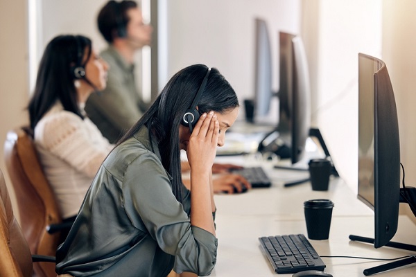 Fotografia de uma jovem agente de telemarketing com mãos na cabeça, que está abaixada, em sinal de estresse. Ela está sentada em frente a uma mesa com um computador em um escritório com outras pessoas ao fundo em atendimento de telemarketing.